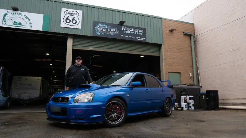 Riley Matthews with a blue WRX STI out front of the RJM Performance workshop, under the RJM sign
