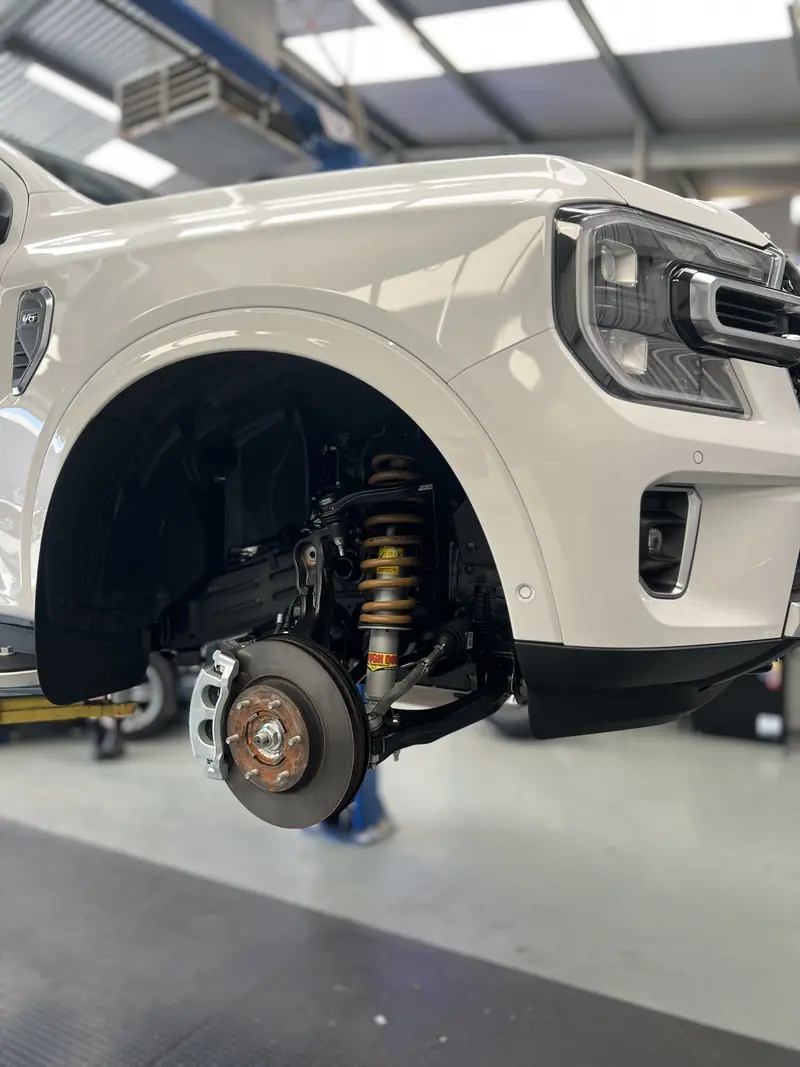 White dual-cab ute on a hoist with the wheel off, showing a gold coilover and brake rotor